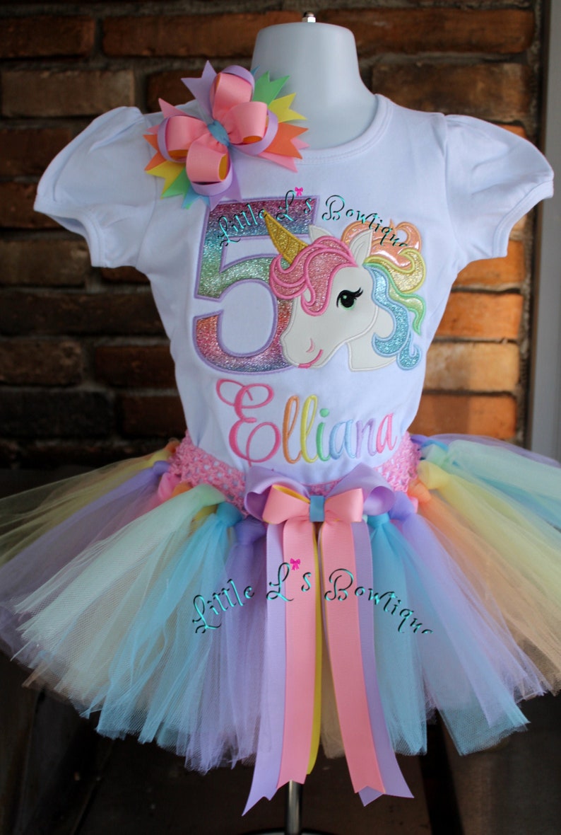May include: A white t-shirt with a rainbow unicorn and the number 5, the name "Elliana" and the text "Little L's Boutique". The shirt is paired with a rainbow tulle tutu with a pink bow.