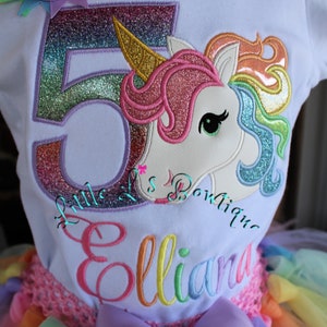 May include: A white t-shirt with a rainbow unicorn and the number 5 embroidered on it. The shirt also has the name "Elliana" embroidered on it. The shirt is paired with a rainbow tulle skirt.