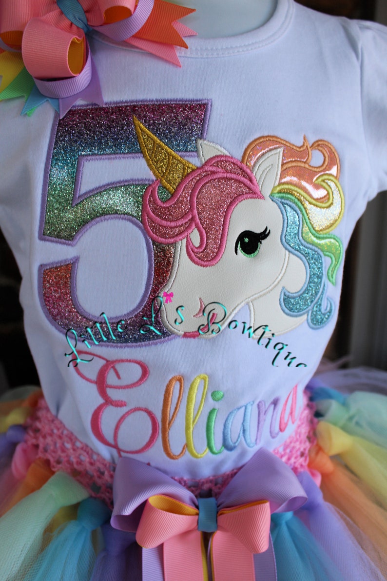 May include: A white t-shirt with a rainbow unicorn and the number 5 embroidered on it. The shirt also has the name "Elliana" embroidered on it. The shirt is paired with a rainbow tulle skirt.