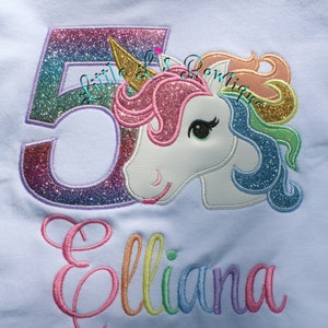 May include: A light blue t-shirt with a rainbow glitter number 5 and a rainbow glitter unicorn with the name "Elliana" embroidered in rainbow colors.