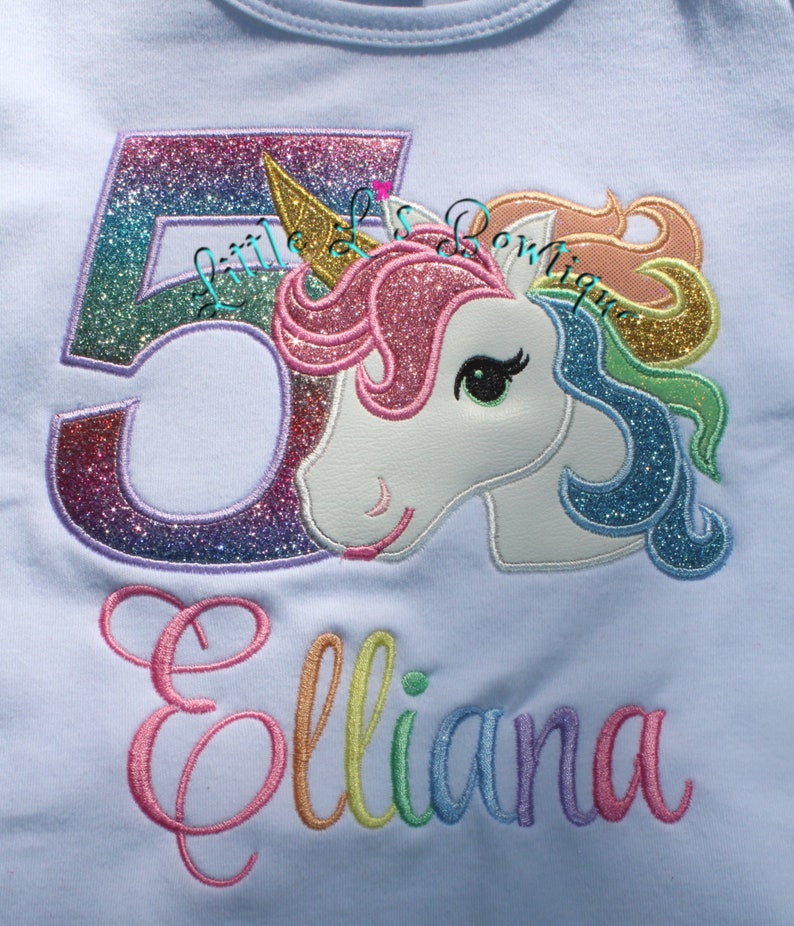 May include: A light blue t-shirt with a rainbow glitter number 5 and a rainbow glitter unicorn with the name "Elliana" embroidered in rainbow colors.