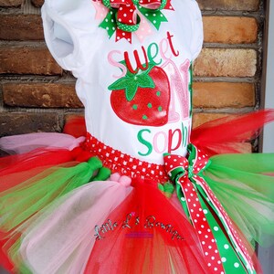 May include: A white onesie with the text "Sweet One" and a red strawberry graphic. The onesie is paired with a multi-layered tulle skirt in pink, red, and green. The skirt has a red and white polka dot ribbon bow.
