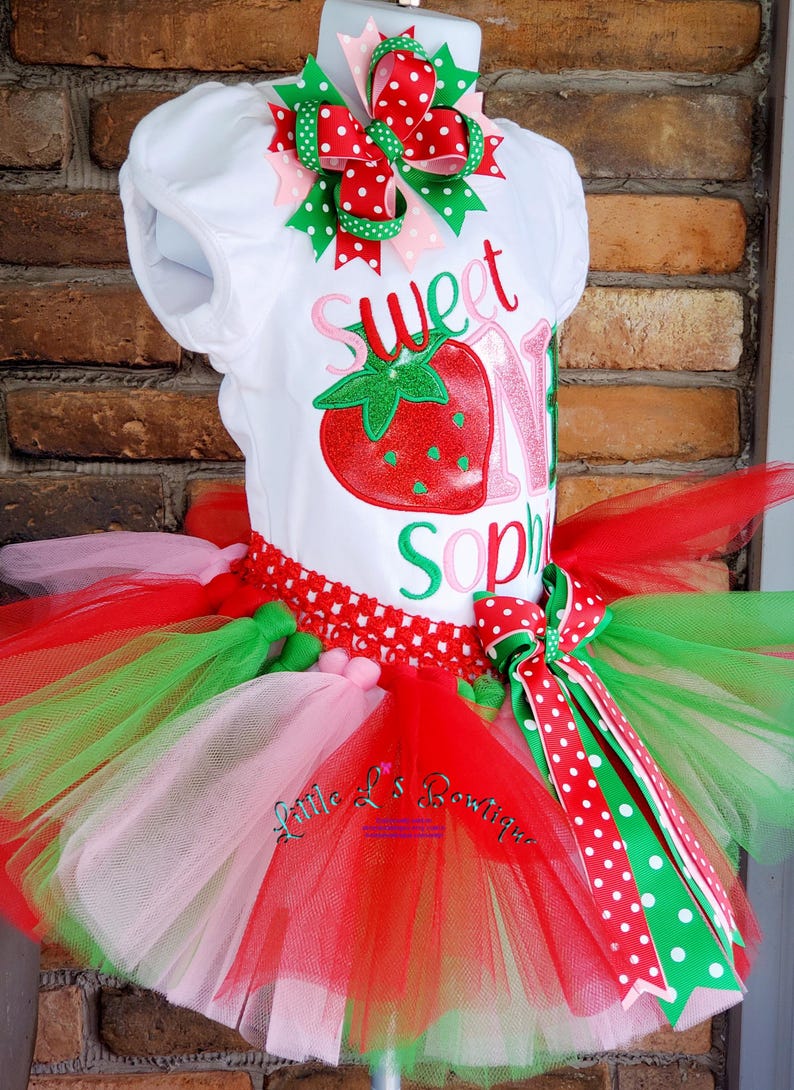 May include: A white onesie with the text "Sweet One" and a red strawberry graphic. The onesie is paired with a multi-layered tulle skirt in pink, red, and green. The skirt has a red and white polka dot ribbon bow.