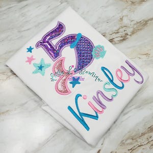 May include: White t-shirt with embroidered mermaid-themed design. Features a large number 59 in purple and pink sequins, a mermaid tail, stars, and the name "Kinsley" in blue, pink, and purple. The shirt is folded.