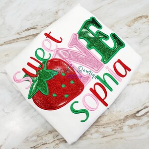 May include: White t-shirt with red, green, and pink embroidered text that says "Sweet One" with a red strawberry graphic and the name "Sophia" embroidered below.