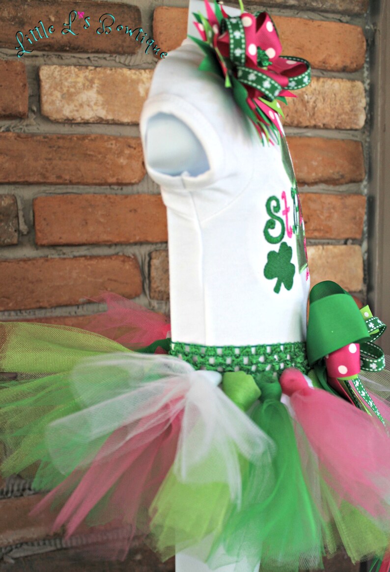Peut inclure: Un body blanc avec un tr&egrave;fle vert et le texte "St. Patty's Day" en rose et vert. Le body est associ&eacute; &agrave; un tutu en tulle vert, rose et blanc avec un n&oelig;ud vert et rose.