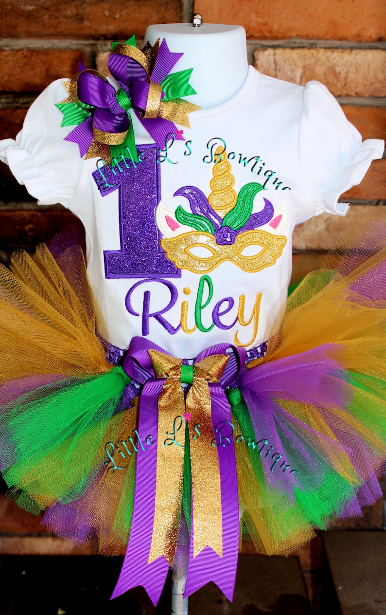 May include: A white onesie with a purple glittery number one and a gold glitter unicorn mask with green leaves. The onesie also has the name Riley embroidered in green. The onesie is paired with a gold, purple, and green tulle tutu with a purple and gold bow. The tutu has the words 'Little L's Boutique' embroidered on the bow.
