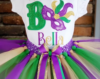 May include: A white shirt with a green letter "B" and a purple mask with gold glitter. The shirt also has the name "Bella" embroidered in gold glitter. The shirt is paired with a multi-layered tulle skirt in green, purple, and gold.