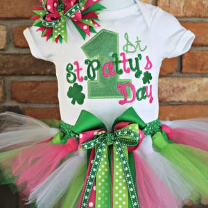 Peut inclure: Un body blanc brod&eacute; de vert et de rose avec l'inscription "1st St. Patty's Day". Le body est associ&eacute; &agrave; un tutu en tulle vert, blanc et rose avec un n&oelig;ud &agrave; pois vert et rose.