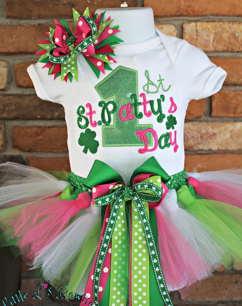 Peut inclure: Un body blanc brod&eacute; de vert et de rose avec l'inscription "1st St. Patty's Day". Le body est associ&eacute; &agrave; un tutu en tulle vert, blanc et rose avec un n&oelig;ud &agrave; pois vert et rose.