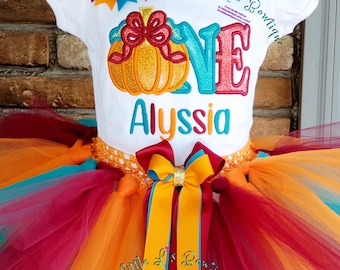 May include: A white short-sleeved shirt featuring an embroidered orange pumpkin, the word ONE, and the name Alyssia in teal. A multi-colored bow sits at the neckline. A layered tulle tutu in orange, teal, and maroon features a matching bow at the waist.