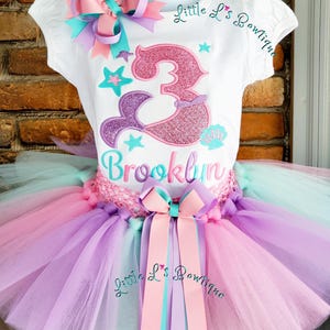 May include: A white t-shirt with a mermaid-themed design, featuring a glittery number 3 and the name "Brooklyn". The outfit includes a multi-colored tutu skirt in shades of pink, purple, and teal, and is adorned with bows.