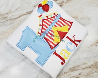 May include: White birthday shirt with a large blue number 1, red and white striped gift boxes, and a cluster of balloons. The name "Jack" is embroidered in red and yellow. The shirt is a festive design for a first birthday.