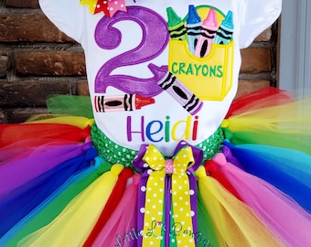 Puede incluir: Un atuendo colorido con una camiseta blanca con el número 2 morado y gráficos de crayones, combinado con una falda tutú arcoíris. La camiseta tiene un lazo y las palabras "Crayons" y "Heidi". El tutú está hecho de capas de tul.