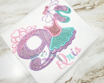 May include: A white t-shirt with a purple glittery number 9, a pink crown, and a ballerina girl in a pink and teal tutu. The text "Little L's Boutique" is embroidered on the shirt.