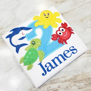 May include: A white t-shirt featuring an embroidered light blue number two, the name James in blue, and colorful sea creature appliques including a yellow octopus, a red crab, a green sea turtle, and a blue shark with white accents.