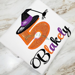 May include: A white t-shirt with an orange glitter number 5, a black witch hat with a purple band, and a black spider. The name "Blakeley" is embroidered in black with a purple outline.