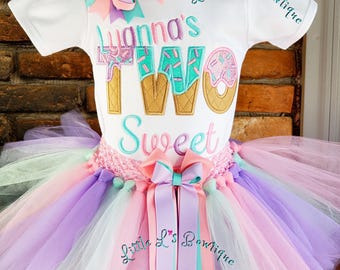 May include: A white toddler's outfit with a tutu skirt. The top has the text "Luanna's TWO Sweet" embroidered in teal, pink, and gold. A large bow and ribbons in pastel colors adorn the outfit. The words "Little L's Boutique" are embroidered on the sleeves.