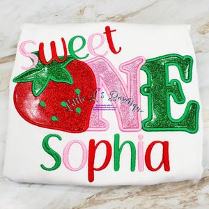May include: A white onesie with the words "Sweet One" in red, pink, and green glitter letters. A red and green glitter strawberry is to the left of the text. The name "Sophia" is written in red, pink, and green glitter letters below the text.