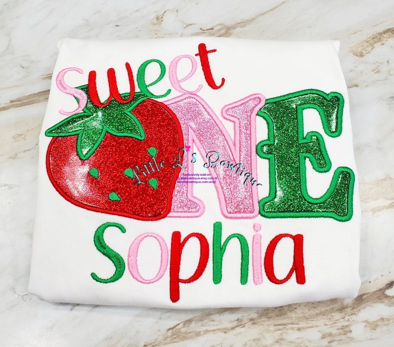 May include: A white onesie with the words "Sweet One" in red, pink, and green glitter letters. A red and green glitter strawberry is to the left of the text. The name "Sophia" is written in red, pink, and green glitter letters below the text.