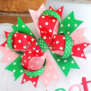 May include: A red, green, and pink polka dot hair bow. The bow is made of three different ribbons, each with white polka dots on a different colored background. The bow is tied in a knot and has a loop on the top.