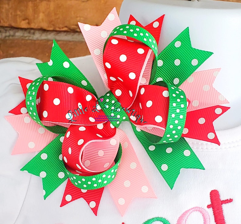 May include: A red, green, and pink polka dot hair bow. The bow is made of three different ribbons, each with white polka dots on a different colored background. The bow is tied in a knot and has a loop on the top.