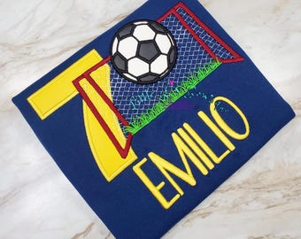 May include: A navy blue t-shirt with a yellow number 7, a red soccer goal, a black and white soccer ball, and the name "EMILIO" in yellow.