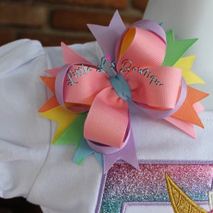 May include: A white shirt with a rainbow unicorn applique and a multi-colored bow with the text "Little L's Boutique" on it.