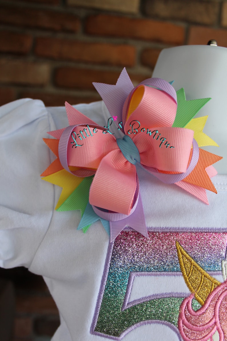 May include: A white shirt with a rainbow unicorn applique and a multi-colored bow with the text "Little L's Boutique" on it.