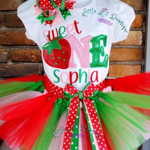 May include: A white t-shirt with the words "sweet one" and a strawberry graphic. The shirt is paired with a red, green, and pink tulle tutu with a red and white polka dot bow.