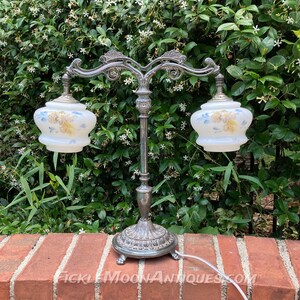 May include: An antique silver-toned table lamp with two frosted glass shades. Each shade features a floral design in yellow and blue. The lamp has a decorative base and a curved arm. The text "FICKLEMOONANTIQUES.COM" is visible on the brick surface.