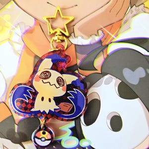 Puede incluir: Un charm de Mimikyu con un cierre dorado en forma de estrella y un charm de Poké Ball. El charm de Mimikyu presenta un patrón a cuadros rojos y negros, detalles azules y una cara amarilla. El fondo presenta personajes de dibujos animados.