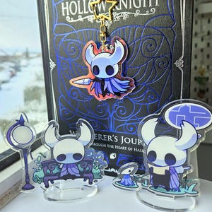 Puede incluir: Una colección de merchandising de Hollow Knight. Incluye un llavero, figuras acrílicas y un libro. El llavero presenta un personaje con una espada. Las figuras representan al personaje en varias poses. El libro tiene el título "Hollow Knight".