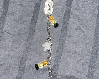 Op de afbeelding: Een zilveren ketting met zwarte en gele kralen, een witte ster-hanger en een spook-hanger die een oranje hoed draagt. Het spook houdt een bezem vast en heeft een paarse ster op zijn hoed.