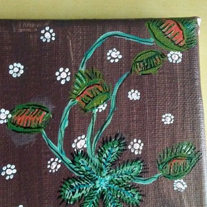 May include: A brown canvas painting featuring a green plant with red and green flowers. The background is brown with white dots.