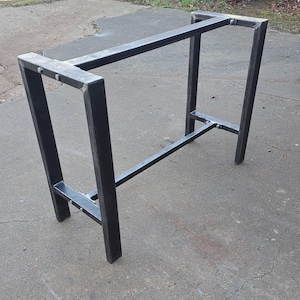 May include: A black metal table base with a rectangular top and a single support beam. The base is made of thick, sturdy metal and has a simple, industrial design.