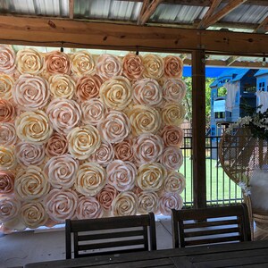 May include: A large paper flower backdrop made of cream and peach colored roses. The backdrop is hanging in a covered outdoor space with a wicker chair and greenery in the background.