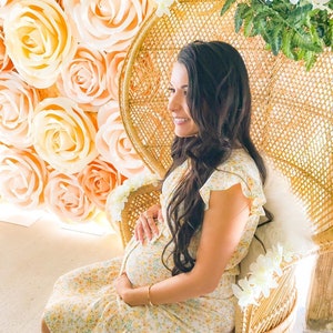 May include: A pregnant person sits in a wicker chair with a floral pattern. The chair is decorated with white flowers. The person is wearing a yellow floral dress and has long brown hair. The background is a wall of large, peach-colored paper roses.