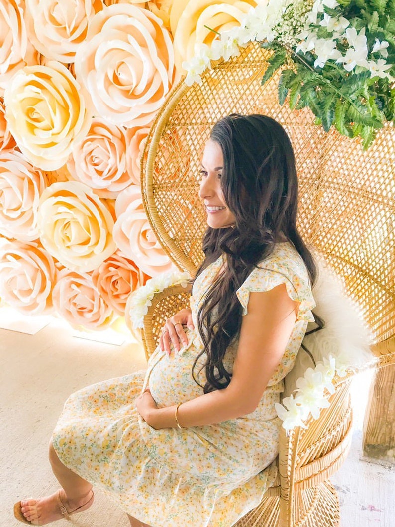 May include: A pregnant person sits in a wicker chair with a floral pattern. The chair is decorated with white flowers. The person is wearing a yellow floral dress and has long brown hair. The background is a wall of large, peach-colored paper roses.