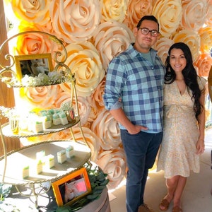 May include: A couple poses in front of a large wall of peach and cream paper roses. The woman is wearing a yellow floral dress and the man is wearing a blue plaid shirt and jeans. They are standing in front of a gold metal tiered stand with small gifts on each tier.