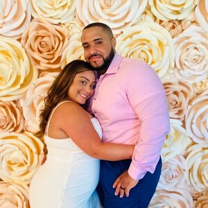 May include: A couple poses for a photo in front of a wall decorated with large, paper, peach and white roses.