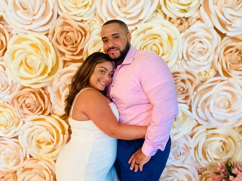 May include: A couple poses for a photo in front of a wall decorated with large, paper, peach and white roses.