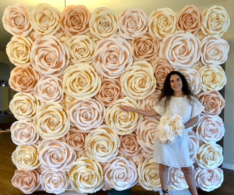 May include: A large wall backdrop made of paper flowers in shades of cream and peach. The flowers are arranged in a repeating pattern and create a romantic and elegant look.