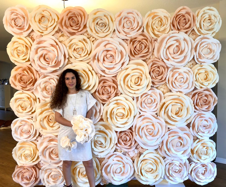 May include: A large wall of paper flowers in shades of cream and blush. The flowers are arranged in a grid pattern and create a beautiful backdrop for a photo shoot or event.