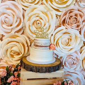 May include: A two-tiered white wedding cake with a brown layer in the middle, decorated with a gold glitter topper that says "Mr. & Mrs. Roman". The cake is on a wooden cake stand and in front of a wall of large, paper, cream-colored roses.