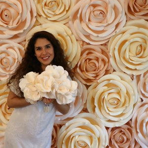 May include: A woman in a white dress holds a bouquet of white paper flowers in front of a wall of large, peach-colored paper roses.