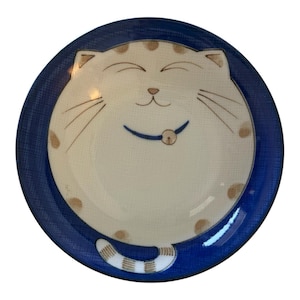 May include: A round ceramic plate featuring a cream-colored cat illustration with closed eyes, a small nose, whiskers, and a blue collar with a bell. The cat has light brown spots and a striped tail. The plate has a wide, textured dark blue rim.