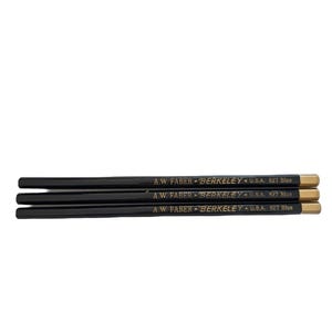 May include: Three black pencils with gold-colored ends are arranged horizontally. The pencils have the text "A.W. Faber Berkeley U.S.A. 827 Blue" printed on them. The pencils are set against a white background.