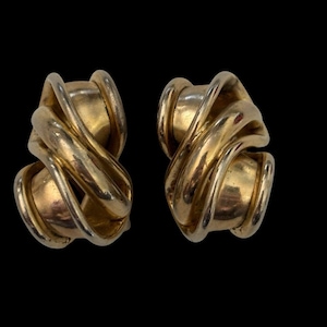 May include: A pair of gold-toned clip-on earrings. Each earring features a central, rounded element wrapped with a swirling, textured design. The earrings are set against a black background.