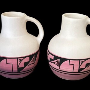 May include: Two white ceramic pitchers with pink and black geometric designs. The pitchers have a rounded body and a handle.